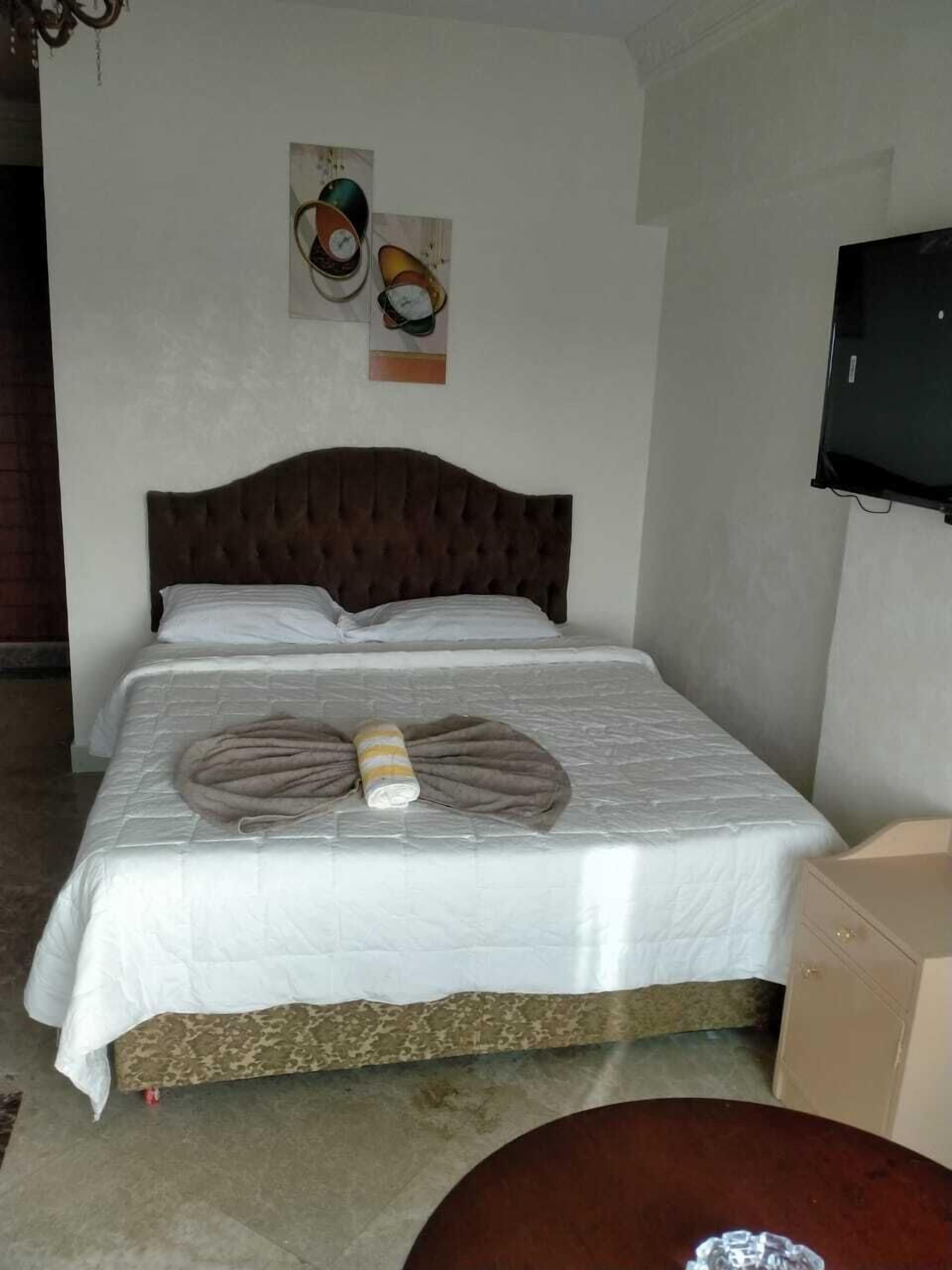 A single bed with a brown headboard, white bedding, and a decorative pillow arrangement. A television is mounted on the wall to the right.
