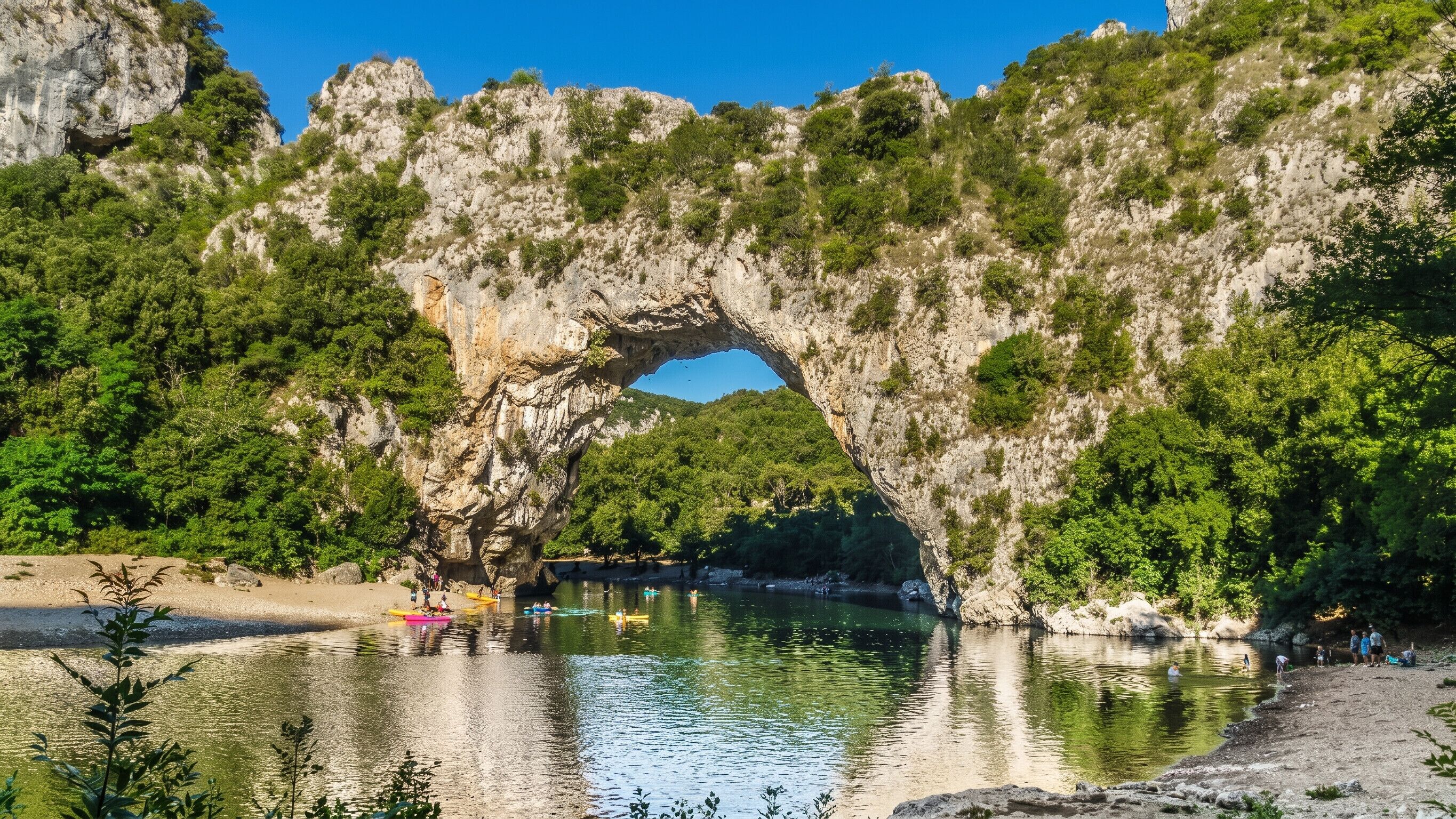 A natural rock arch over a calm river with people kayaking.