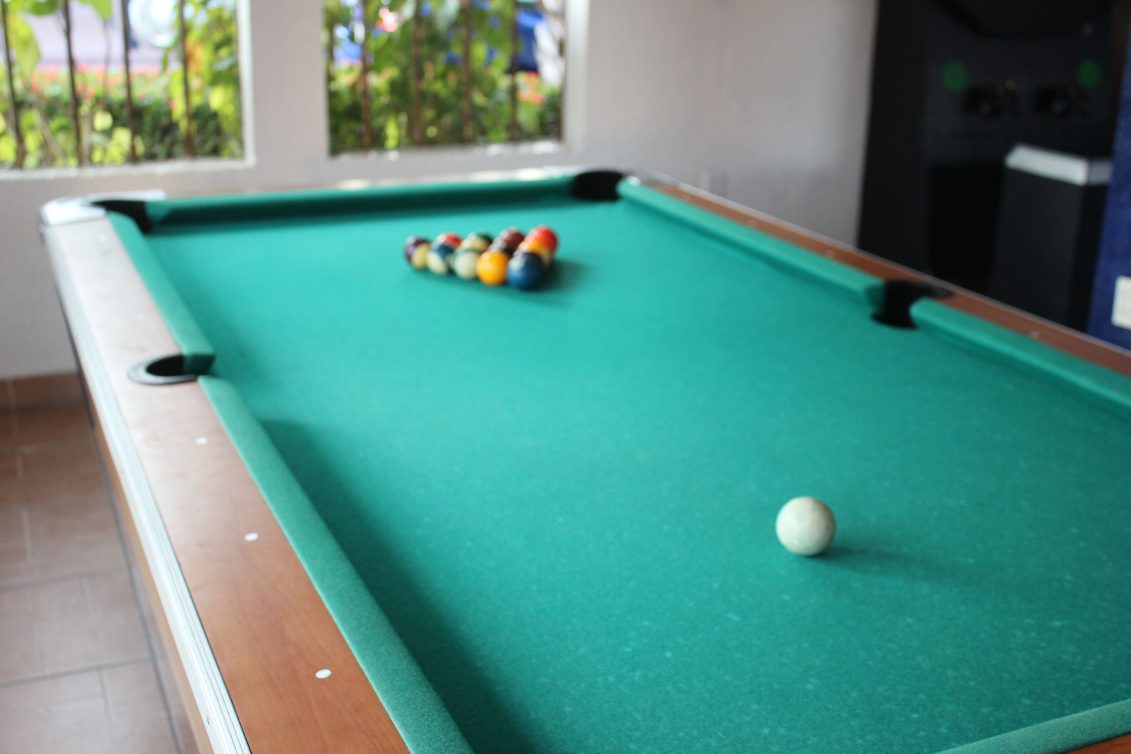 A pool table with balls arranged in a triangle formation, a white cue ball, and a window with a view of greenery.