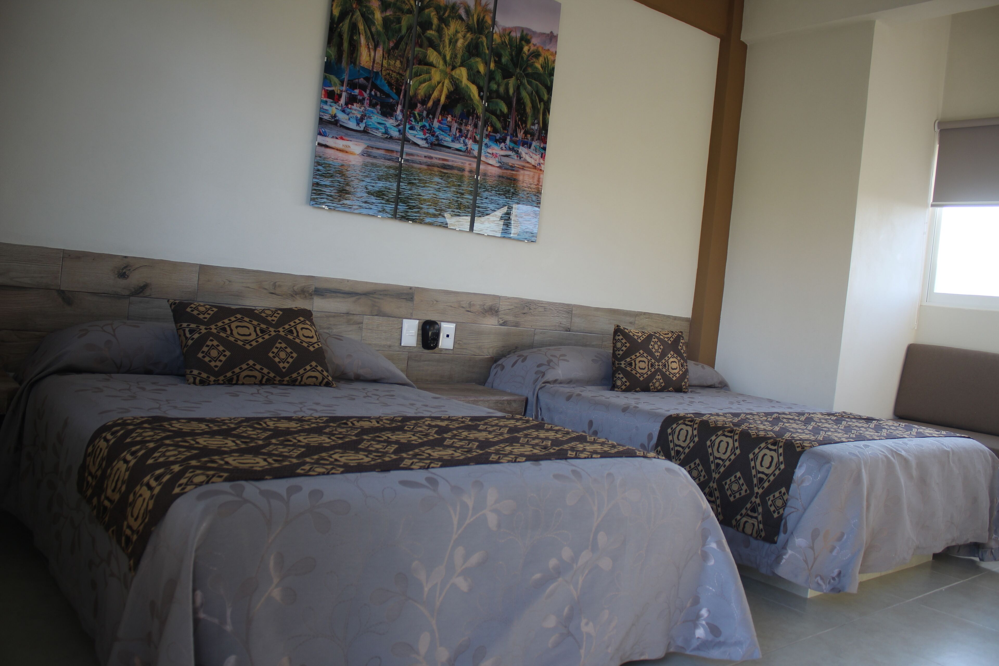 Habitación de hotel con dos camas, cabecero de madera, pintura mural de una playa y ventana con vistas a palmeras.