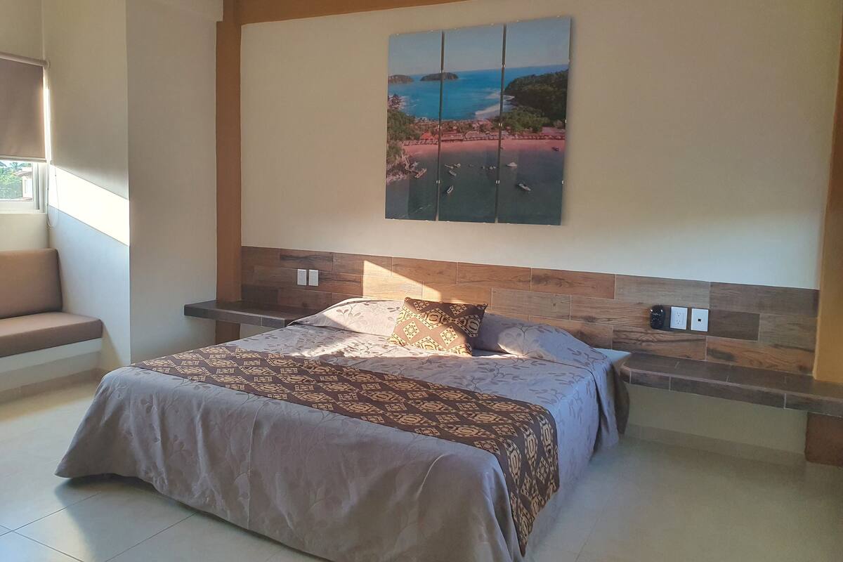A bedroom with a large bed, a nightstand, a bench, and a wall-mounted painting of a coastal scene.