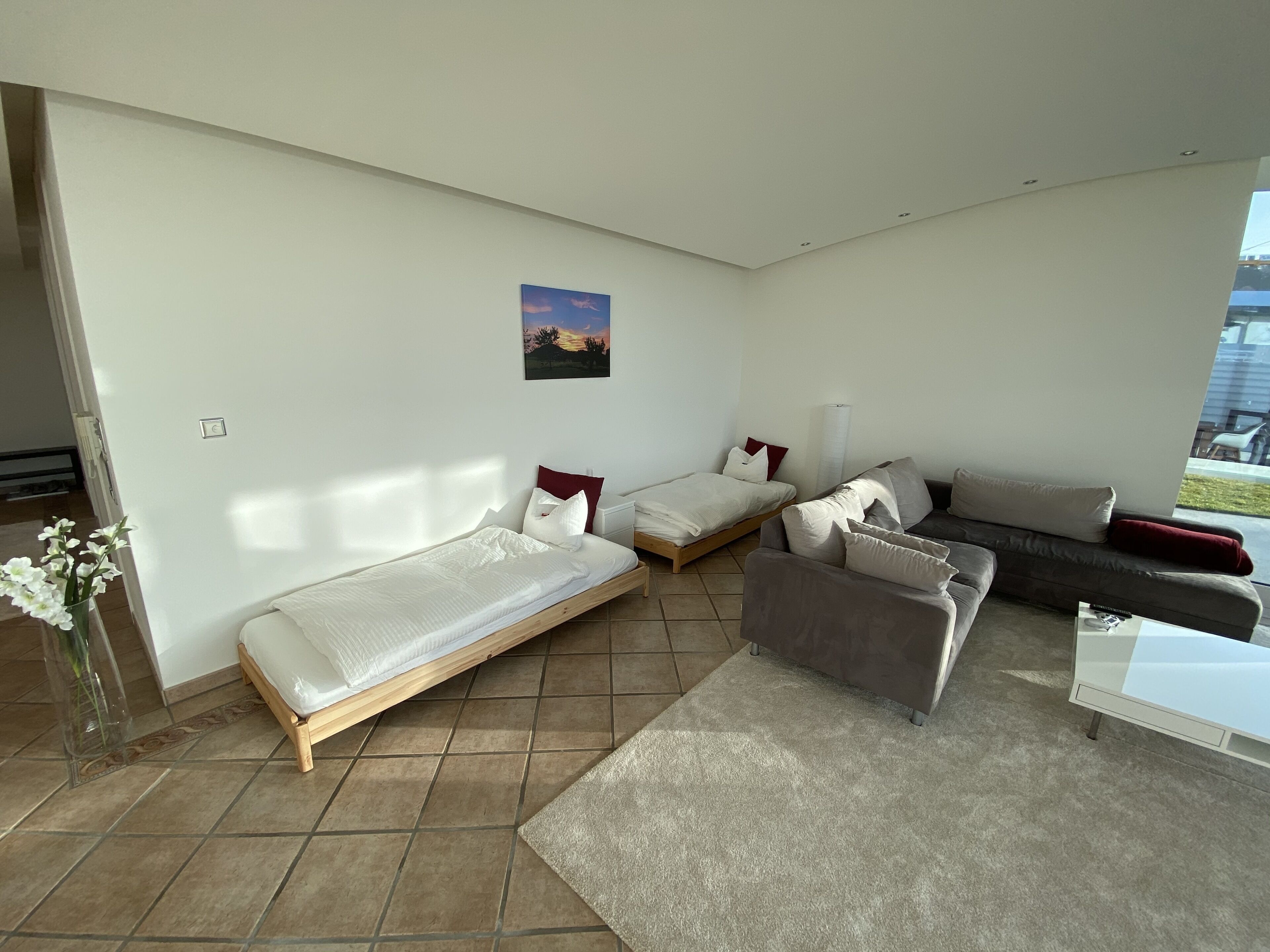 A modern, well-lit room with a large window, a sofa, two single beds, and a small table.