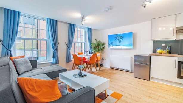 A modern living room with a grey sofa, orange cushions, a white coffee table, and a dining area with orange chairs. There is a kitchen with white cabinets and a small refrigerator. - London Street, Andover, Hampshire, SP102NU, United Kingdom (Andover)