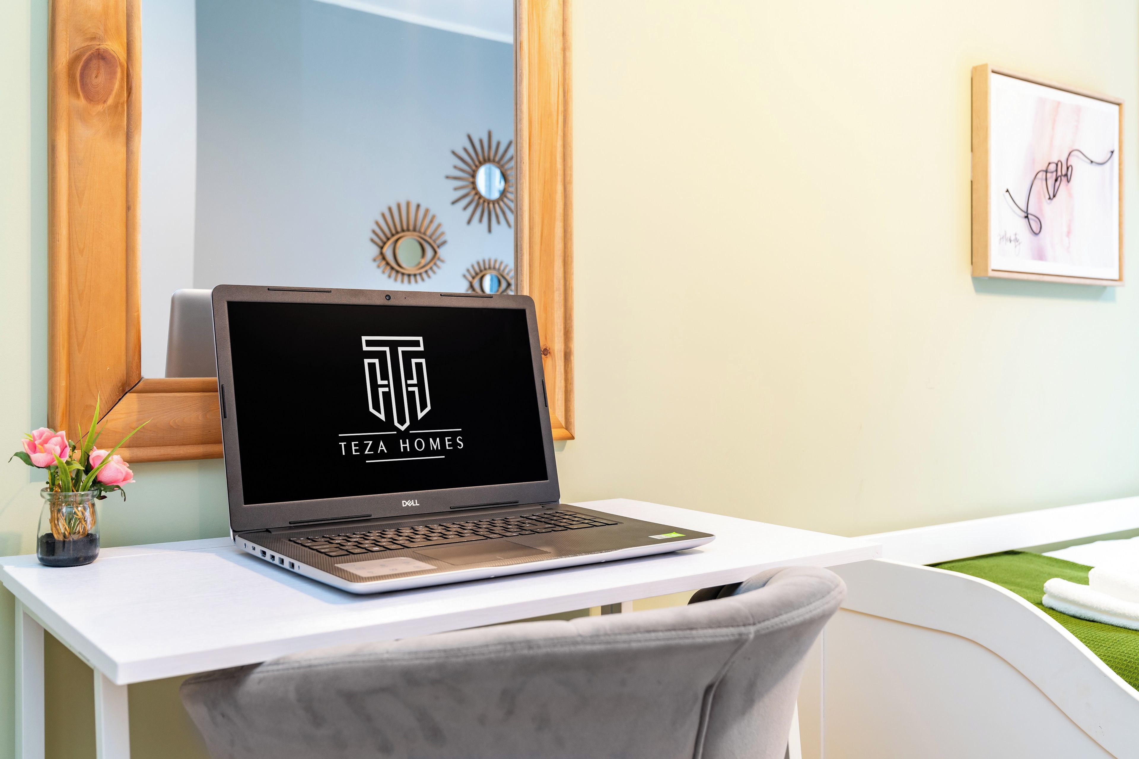 A laptop on a white desk with a vase of flowers, a mirror, and wall art.