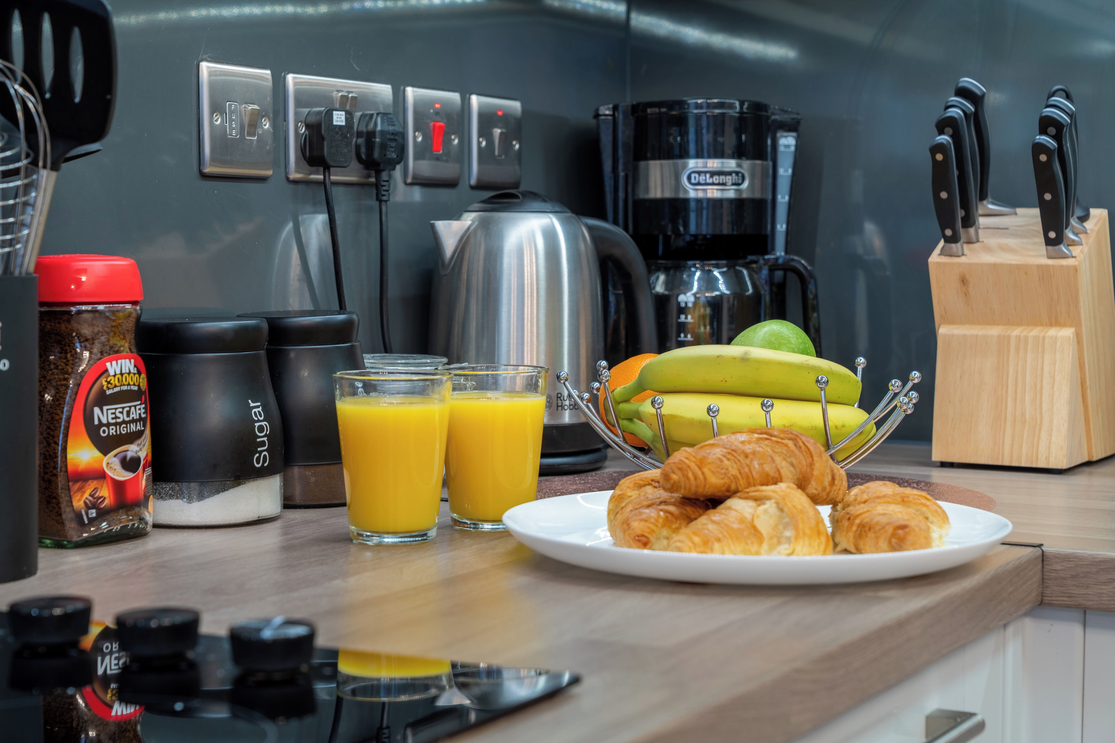 En köksbänk med en kaffebryggare, en vattenkokare, ett knivställ, en tallrik med croissanter och två glas apelsinjuice.