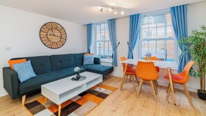 A modern living room with a dark blue sofa, white coffee table, and orange chairs around a white table. A large wall clock and a potted plant are visible. - London Street, Andover, Hampshire, SP102NU, United Kingdom (Andover)