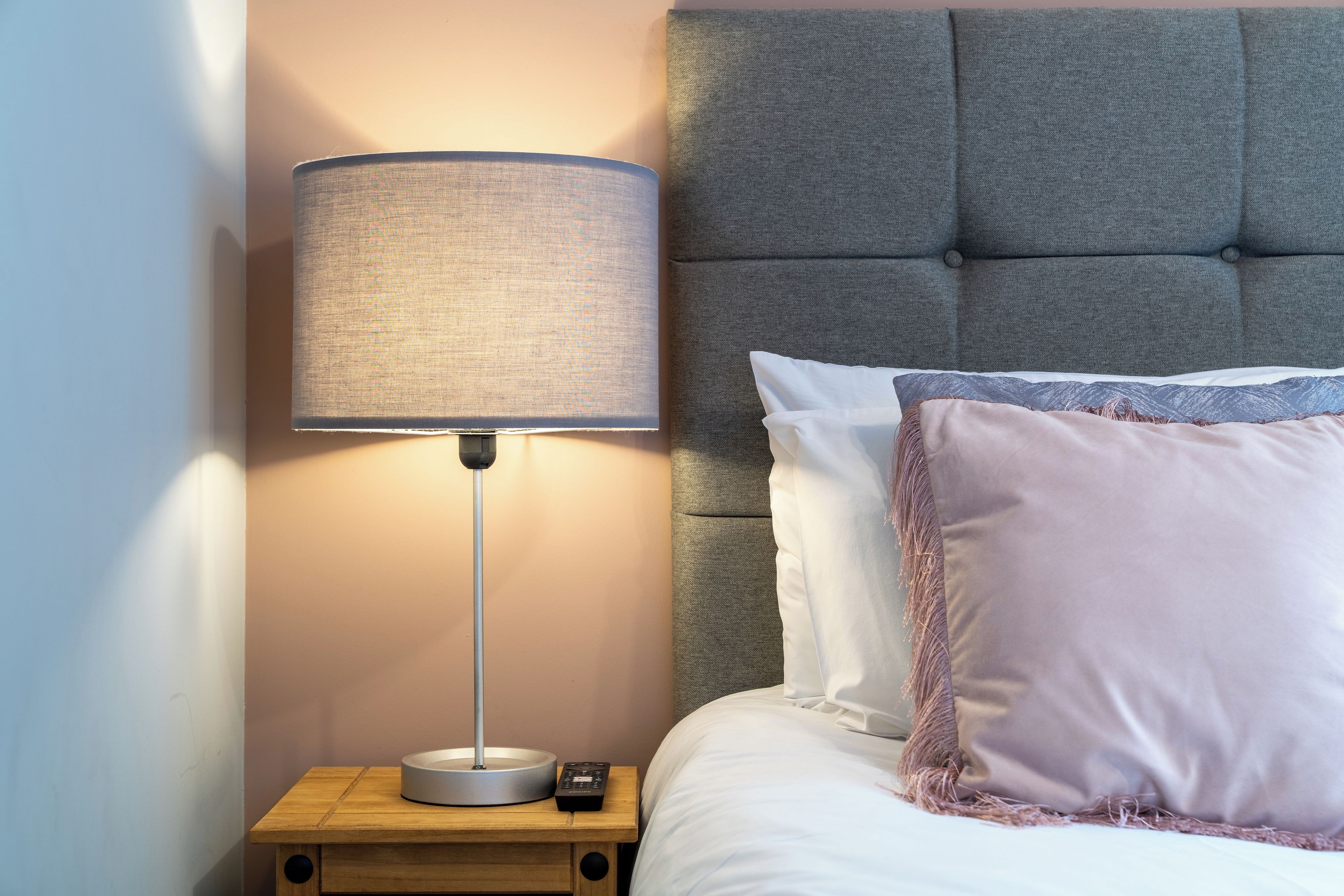 Una cama bien hecha con cabecera gris, una mesita de noche con lámpara y control remoto.