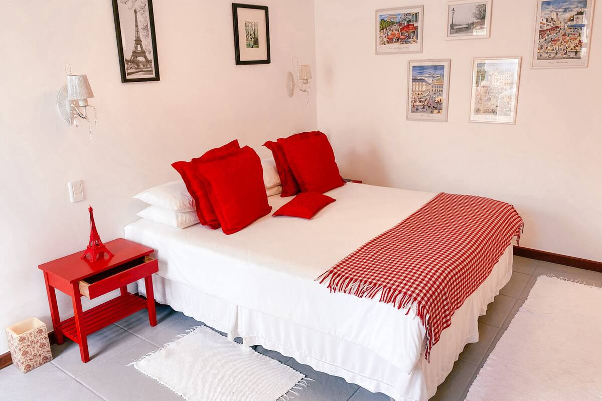 Un dormitorio con una cama blanca, almohadas rojas, una mesita de noche roja y una pequeña reproducción de la Torre Eiffel.