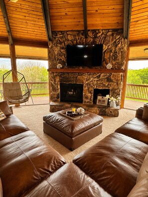 Un área de estar exterior acogedora con chimenea de piedra, asientos de cuero y un televisor de pantalla plana montado sobre el hogar.