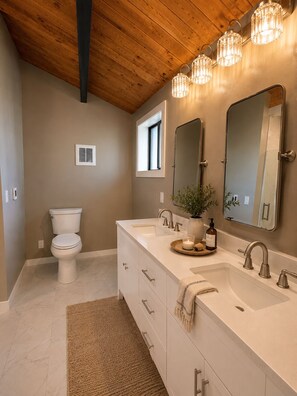 Une salle de bain équipée de toilettes, de deux lavabos, d’un miroir et d’un luminaire au plafond.