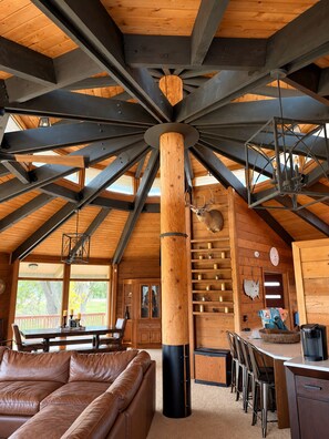 A spacious interior with a high ceiling, wooden beams, a central wooden column, a dining area with chairs and a table, a leather sofa, and a fireplace.
