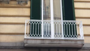 Fachada de edificio con unidades de aire acondicionado, una ventana con contraventanas verdes y un balcón blanco.