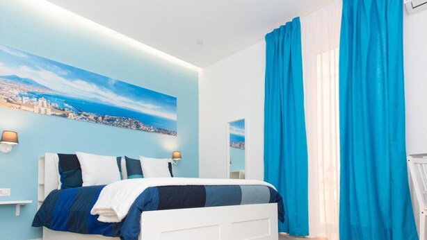 Un dormitorio moderno con un mural de paisaje urbano, una cama blanca y cortinas azul brillante.