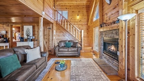 A cozy log cabin interior with a fireplace, leather sofas, a wooden staircase, and a dining area.