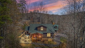 A log cabin with a green roof, surrounded by trees and a gravel driveway.