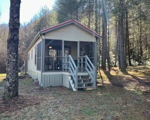 A small cabin with a porch and a staircase, surrounded by trees.