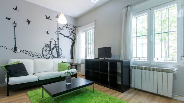 Un salon avec un canapé blanc, des coussins verts, une table basse noire, un téléviseur posé sur un meuble noir et un tapis vert.