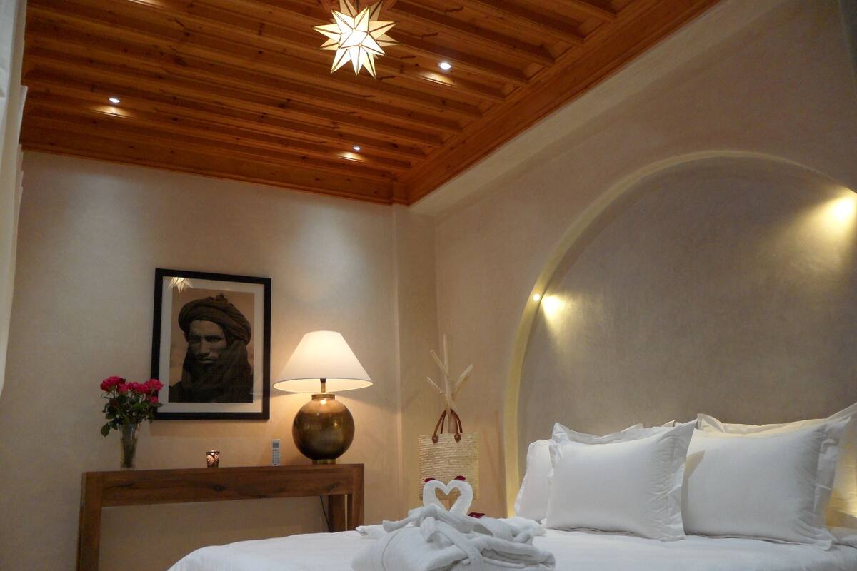 Habitación de hotel con techo de madera, cama con ropa blanca, mesita de noche con lámpara, cuadro en la pared y un jarrón con flores.