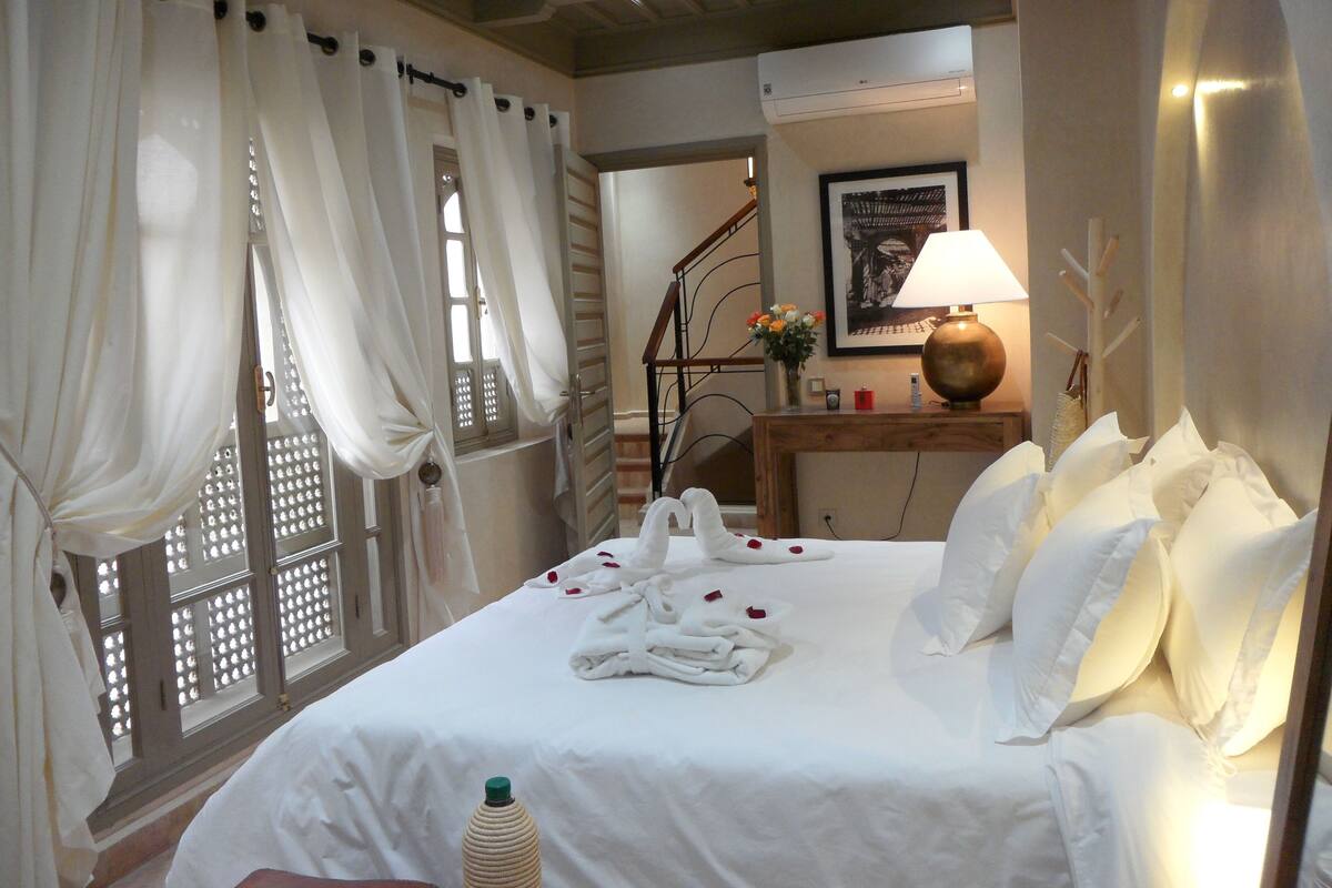 Una habitación de hotel con una cama bien hecha, cortinas blancas, un cabecero de madera, una lámpara de noche y un cuadro en la pared.