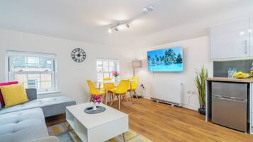 A modern living room with a sofa, coffee table, and a dining area with yellow chairs. A flat-screen TV is mounted on the wall, and there is a kitchen area with a refrigerator.