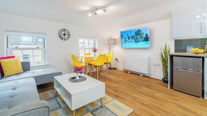 A modern living room with a sofa, coffee table, and yellow chairs. There is a dining table with chairs, a TV mounted on the wall, and a kitchen area with a fridge and cabinets. - London Street, Andover, Hampshire, SP102NU, United Kingdom (Andover)