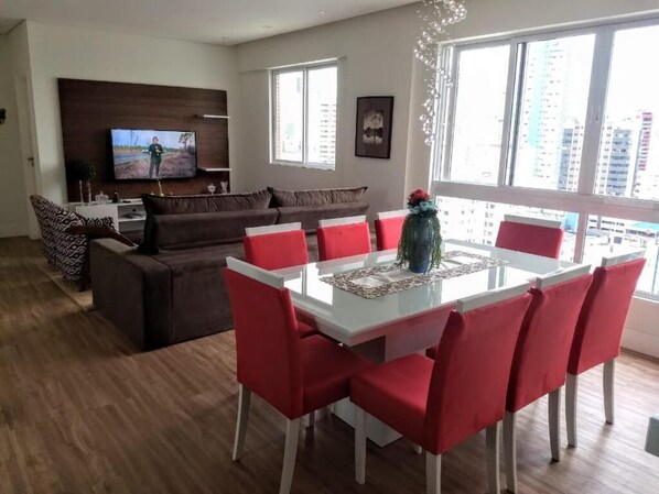 A modern living room with a brown sofa, a white dining table with red chairs, and a flat-screen TV mounted on the wall.