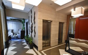A modern lobby with a geometric patterned floor, two elevators, and a seating area with a plant.