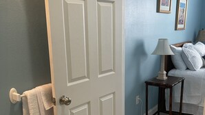 A bathroom with a white door, a towel rack with a towel, a small table with a lamp, and a bed with pillows.