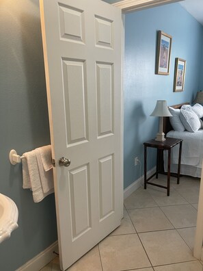 A bathroom with a white door, a towel rack with a towel, a small table with a lamp, and a bed with pillows.