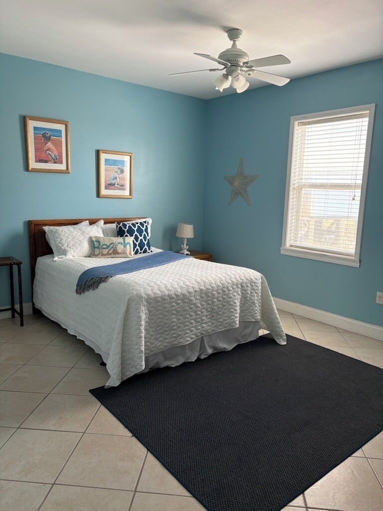 Une chambre à coucher comprenant un lit, un ventilateur de plafond, une fenêtre avec des stores et des éléments de décoration murale.