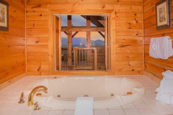A wooden cabin bathroom with a large bathtub, a window with a view of mountains, and a wooden deck outside.