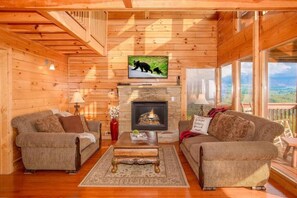 A cozy cabin interior with wooden walls, a fireplace, two sofas, and a television mounted above the fireplace.