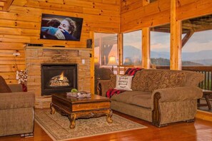 A cozy living room with a fireplace, a television mounted on the wall, and a large window offering a view of the outdoors.