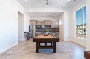 A modern kitchen with a pool table, grey cabinets, and a view of a residential area.