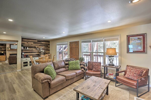A spacious living room with a brown leather sofa, a wooden coffee table, and a brown leather armchair. There is a dining area with a wooden table and chairs, and a kitchen in the background. - Walk to Marina: Rustic Grand Lake Retreat! (Grand Lake)