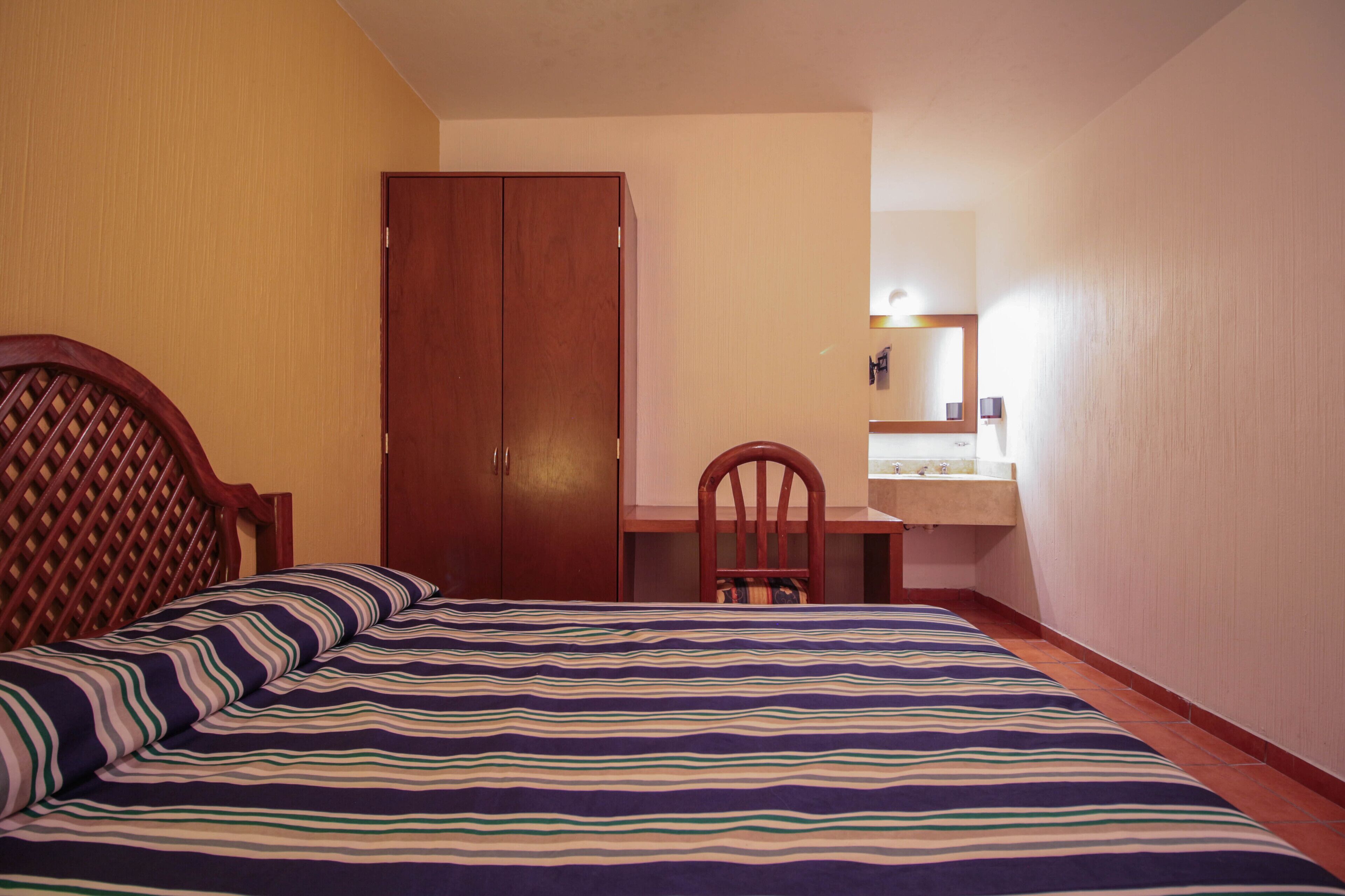 Un dormitorio con cama, armario de madera, escritorio con silla y baño con espejo y lavamanos.