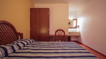 Un dormitorio con cama, armario de madera, escritorio con silla y baño con espejo y lavamanos.