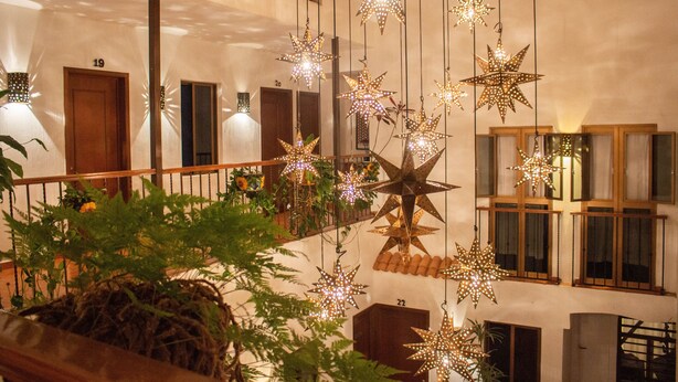 Interior de un hotel con varias habitaciones, luces colgantes en forma de estrella y un balcón con plantas.