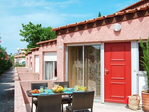 A row of pink buildings with numbered doors, a red door, and a table with chairs and fruit.