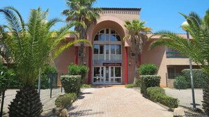 Un bâtiment à la façade rouge, doté d’une entrée en arc de cercle et bordé de palmiers au premier plan.