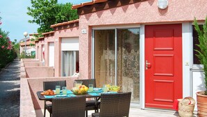 Une rangée de bâtiments roses avec des portes numérotées, une porte rouge et une table entourée de chaises, avec des fruits dessus.