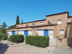 A building with a beige facade, red-tiled roof, and blue doors. There are two garage doors, a small air conditioning unit, and a white fence.