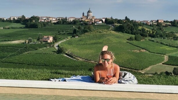Una persona descansando junto a una piscina con vistas a viñedos y un edificio lejano.