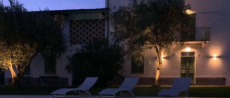 Un área de piscina con sillas reclinables, un edificio con balcón y un árbol en primer plano.