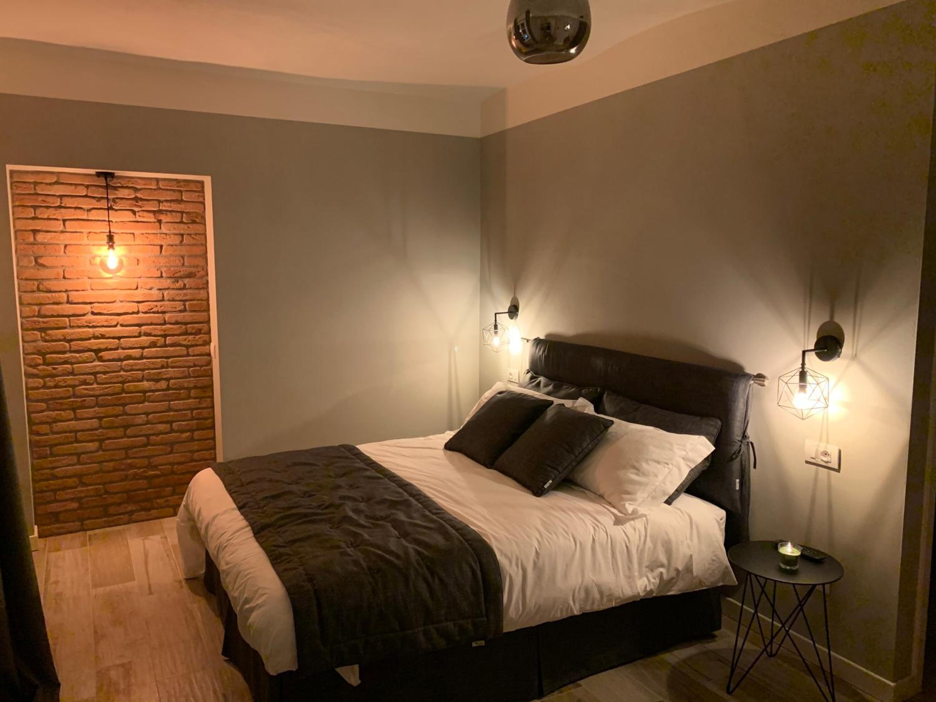 Een slaapkamer met een bakstenen muur, een bed met donker hoofdeinde, twee wandlampen en een klein rond tafeltje met een afstandsbediening.