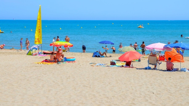 Een strand met mensen die zonnebaden en zwemmen, kleurrijke parasols, en een helderblauwe lucht.