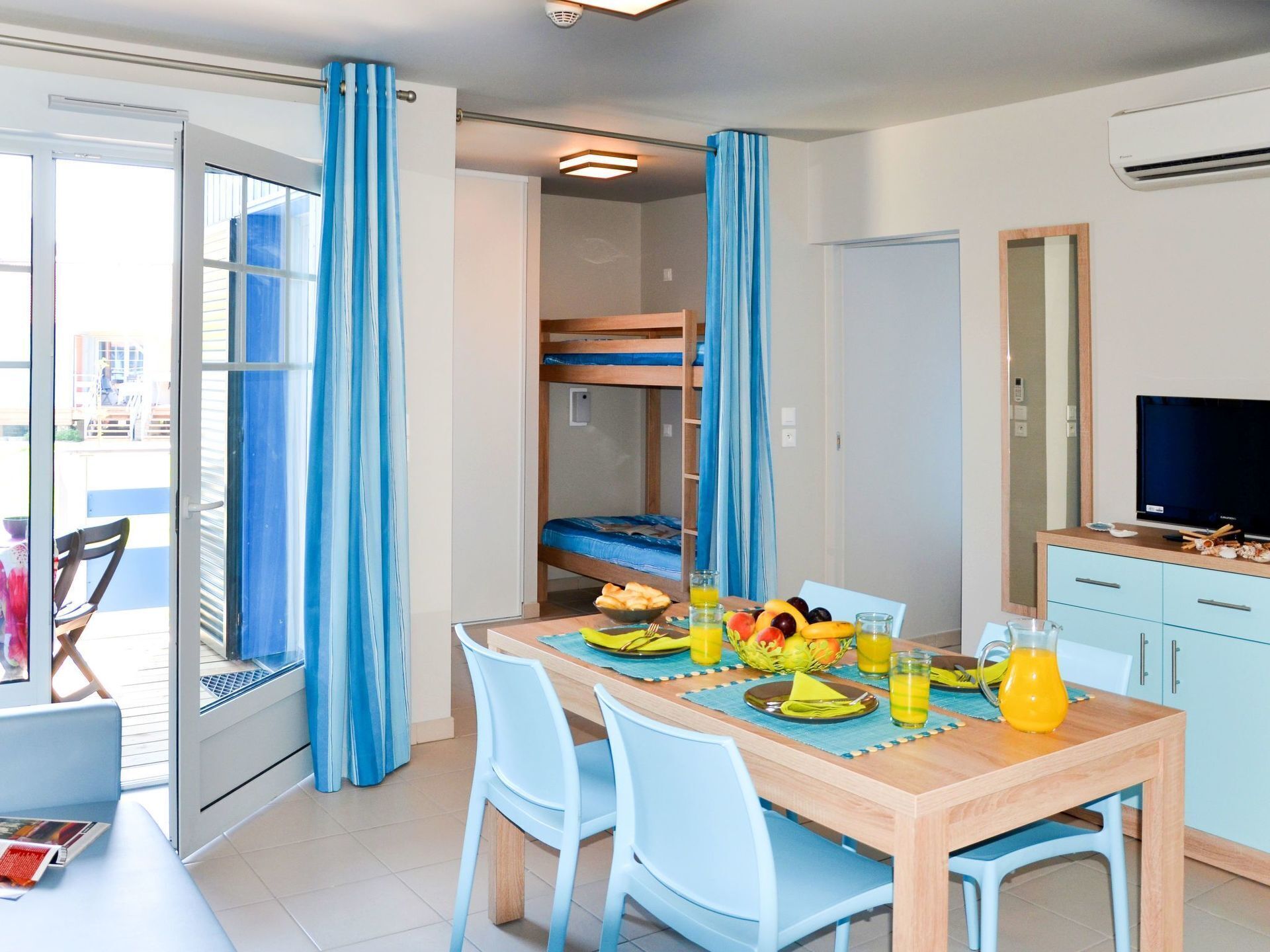 Una stanza con un tavolo da pranzo apparecchiato per due persone, una cuccetta, una televisione e un balcone con tende blu.