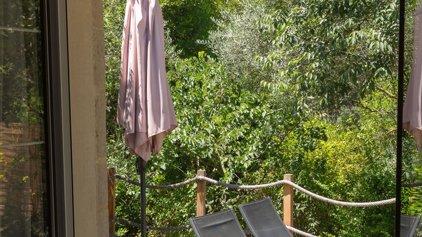 A glass door leading to an outdoor patio with two lounge chairs, a wicker ottoman, and a pink umbrella. - Le Mas des Alexandrins (Uzès)