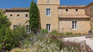 A stone building with multiple windows and a small tower, surrounded by a garden with various plants and flowers. - Le Mas des Alexandrins (Uzès)