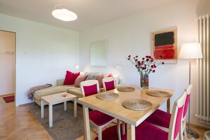 Una sala moderna con una mesa de comedor de madera, sillas tapizadas en rojo, un sofá color beige con cojines rojos, una lámpara de pie y un florero con flores rojas.